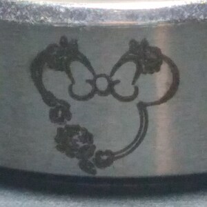 May include: A silver ring with a laser-engraved design of a cartoon mouse with a bow. The mouse is surrounded by a floral design.