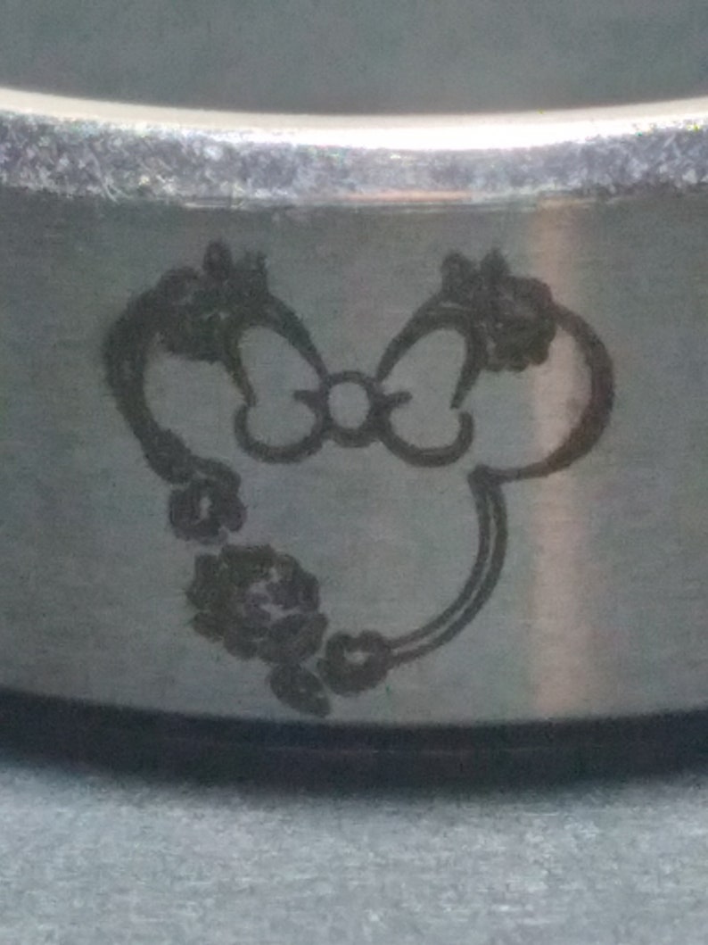 May include: A silver ring with a laser-engraved design of a cartoon mouse with a bow. The mouse is surrounded by a floral design.