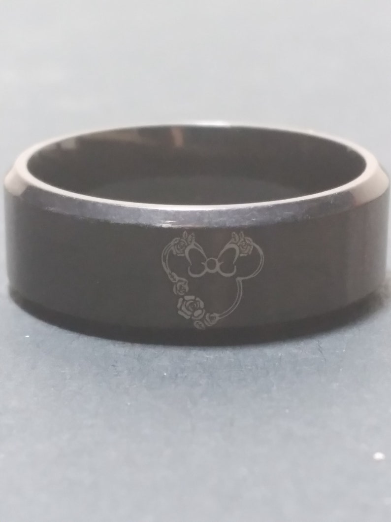 May include: A black stainless steel ring with a laser-engraved design of a cartoon mouse head with a bow and flowers.