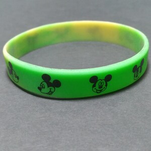 Puede incluir: Una pulsera de silicona verde y amarilla con siluetas negras de la cabeza de Mickey Mouse. La pulsera es de color verde vibrante con un degradado amarillo y tiene forma circular. Las siluetas de Mickey Mouse est&aacute;n espaciadas uniformemente.