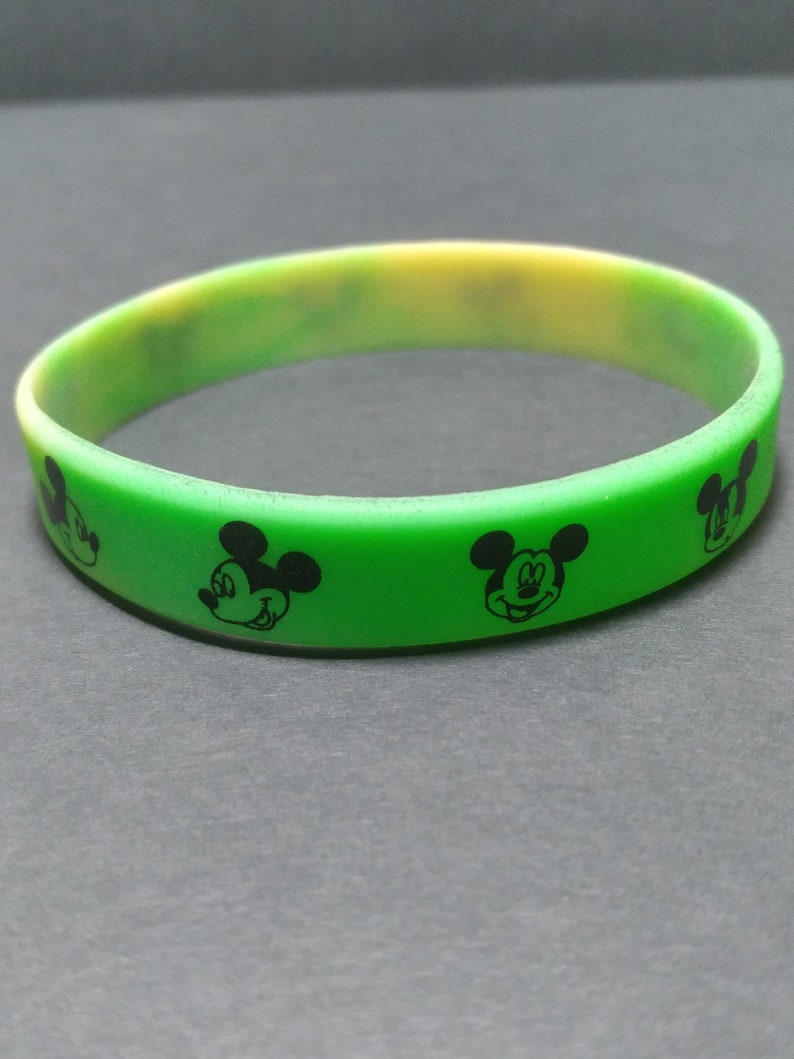 Puede incluir: Una pulsera de silicona verde y amarilla con siluetas negras de la cabeza de Mickey Mouse. La pulsera es de color verde vibrante con un degradado amarillo y tiene forma circular. Las siluetas de Mickey Mouse est&aacute;n espaciadas uniformemente.