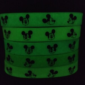 Puede incluir: Una pila de cinco pulseras de silicona verde ne&oacute;n. Cada pulsera presenta un patr&oacute;n repetido de caras negras de Mickey Mouse. Las pulseras brillan intensamente sobre un fondo oscuro.