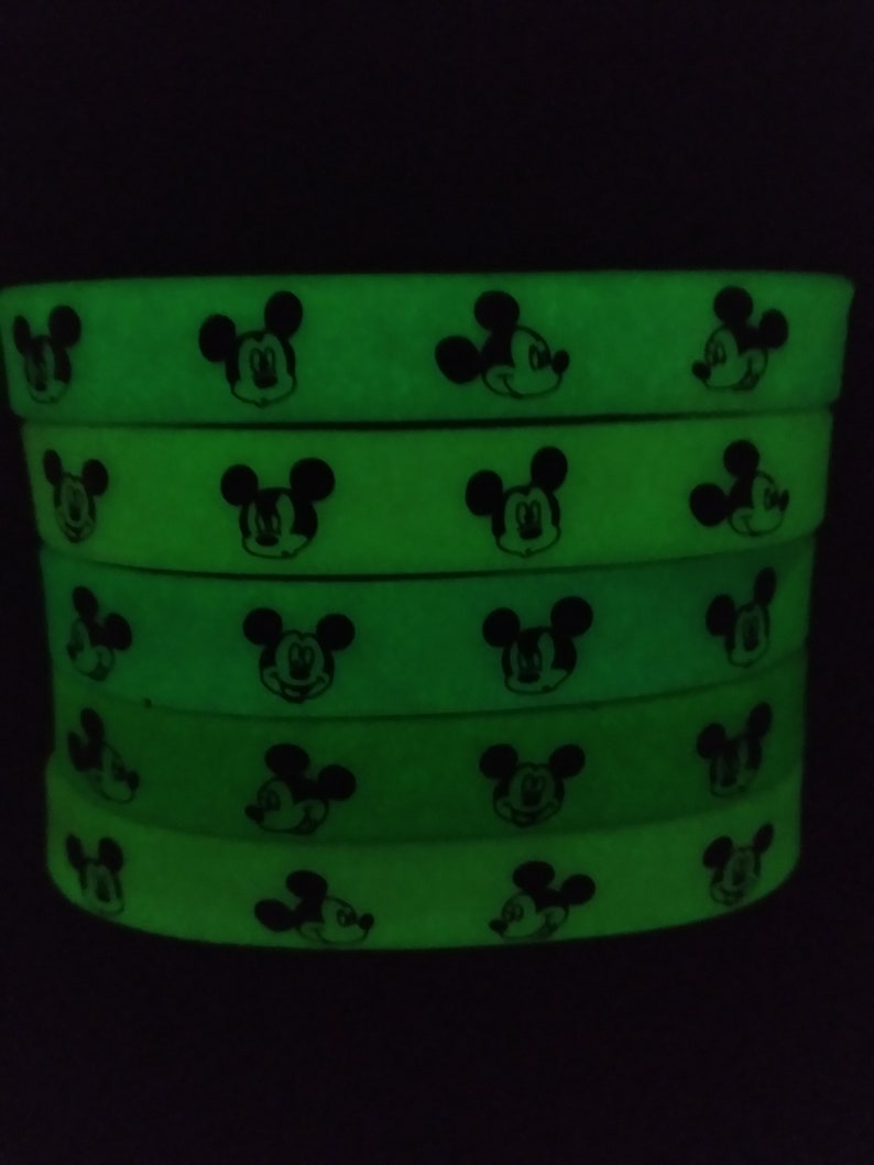 Puede incluir: Una pila de cinco pulseras de silicona verde ne&oacute;n. Cada pulsera presenta un patr&oacute;n repetido de caras negras de Mickey Mouse. Las pulseras brillan intensamente sobre un fondo oscuro.