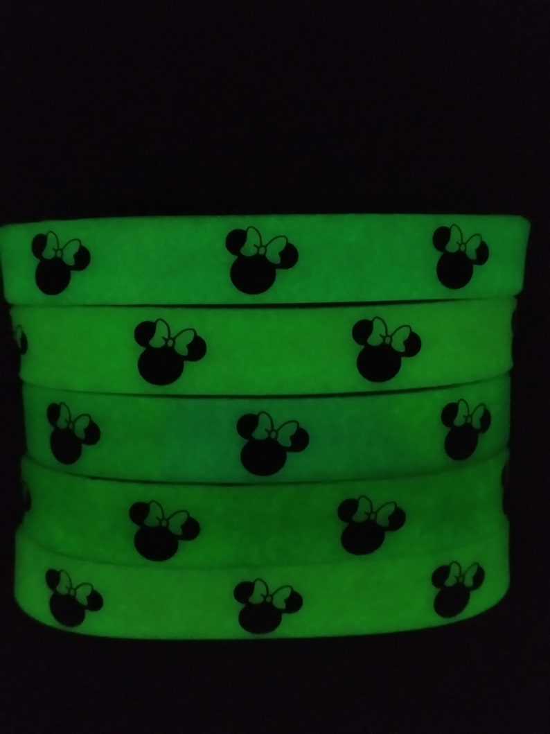 May include: A stack of five glow-in-the-dark green silicone wristbands with a black silhouette of Minnie Mouse's head with a bow on top.