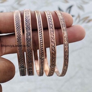 May include: Five copper bangles featuring distinct embossed patterns, including wavy lines, cross-hatching, and diagonal textures. Each bracelet has a metallic finish and the text BhomiyaJewellers is visible on the left side of the image.