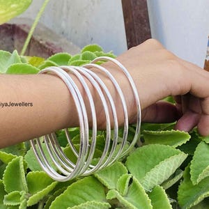 May include: A set of five smooth, polished silver-toned bangles worn on the wrist. The bangles are thin, circular, and have a reflective metallic finish. The text BhomiyaJewellers is visible on the left side of the image near the bangles.