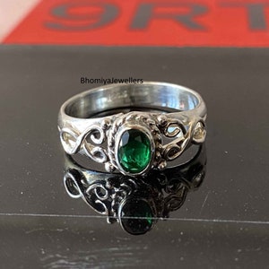 Puede incluir: Un anillo de plata con una piedra preciosa verde engastada en un delicado diseño de filigrana. El anillo está sobre una superficie negra y refleja la luz.