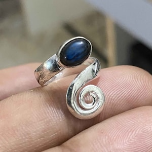 Op de afbeelding: Een zilveren ring met een blauwe ovale edelsteen in een swirl-ontwerp.