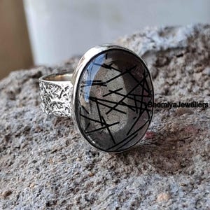 Könnte beinhalten: Silberring mit einem klaren Quarzstein mit schwarzen Einschlüssen. Der Ring hat eine strukturierte Band.