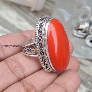 Pode incluir: Um anel marcante com uma grande pedra preciosa vermelha oval. A banda de prata é adornada com padrões decorativos detalhados. Este anel de declaração foi projetado para fazer uma declaração de moda ousada, adicionando um toque vibrante a qualquer roupa.