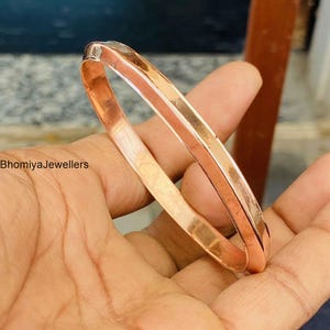 Puede incluir: Una pulsera rígida de cobre pulido con una superficie lisa y reflectante de diseño sencillo y redondeado. El metal tiene un tono metálico cálido, de color naranja rojizo. El texto BhomiyaJewellers es visible a la izquierda de la imagen.