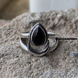 以下が含まれることがあります： 黒い涙型の宝石がついた銀の指輪。三日月模様が施されています。指輪は灰色の岩の上に置かれています。画像の右下には「BhomlyaJewellers」と書かれています。