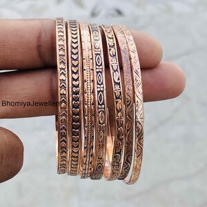 May include: A set of six copper-toned metal bangles featuring various embossed patterns including hearts, geometric shapes, and floral motifs. The text BhomiyaJewellers is visible on the left side of the bangles, which have a metallic finish.