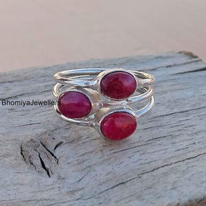 May include: Three silver rings stacked together, each with an oval, deep red gemstone. The rings are displayed on a weathered wooden surface, highlighting the jewellery's design and colour.