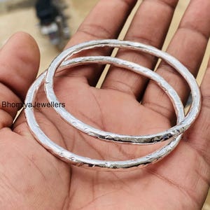 Puede incluir: Dos brazaletes circulares de plata con una superficie martillada y texturizada que refleja la luz. Las bandas de metal tienen un perfil redondeado consistente y un acabado pulido. El texto BhomiyaJewellers es visible en el lado izquierdo.
