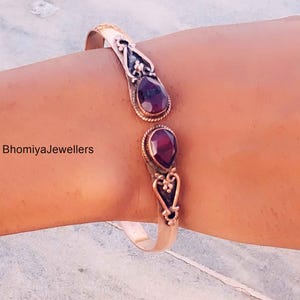 Op de afbeelding: Open manchetarmband met twee druppelvormige, dieprode edelstenen. Het metaal heeft een sierlijk ontwerp met bloemaccenten. De armband wordt om een arm gedragen. De tekst "BhomiyaJewellers" is zichtbaar.