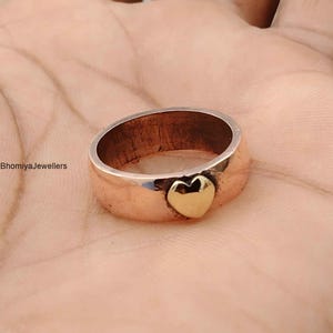 May include: A copper-toned metal ring featuring a smooth, polished surface and a single, small, raised gold-toned heart emblem centered on the band. The text BhomiyaJewellers is visible on the left side of the ring, which has a wide, flat profile.