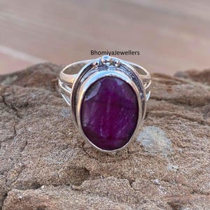 Puede incluir: Un anillo de plata con una gran piedra preciosa ovalada de color morado oscuro engastada en un engaste de bisel. El anillo tiene una banda texturizada con un diseño decorativo.