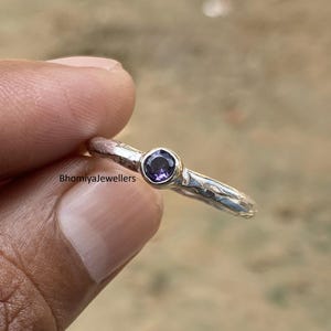 Pode incluir: Um anel de prata com uma pequena pedra preciosa redonda e roxa no centro. A banda do anel tem um aspeto texturizado e martelado. O anel é segurado pelos dedos de uma pessoa. O texto "BhomiyaJewellers" é visível.