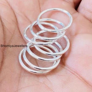 Könnte beinhalten: Ein Stapel aus fünf dünnen, polierten Silberringen mit einem glatten, abgerundeten Banddesign. Die Ringe sind vertikal gestapelt, was ein geschichtetes Aussehen erzeugt. Der Text BhomiyaJewellers ist links neben dem Ringstapel zu sehen.