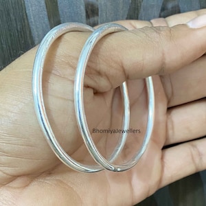 May include: Two silver bangles, one slightly larger than the other, are shown in a close-up image. The bangles are smooth and shiny, with a simple, circular design. The text "Bhomiyajewellers" is visible in the image.
