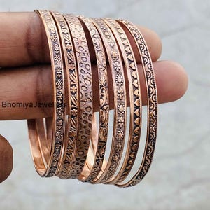 May include: A set of seven copper-toned metal bangles featuring distinct embossed patterns including floral motifs, heart shapes, and textured dots. Each bangle has a metallic finish with darkened recessed details. The text BhomiyaJewellers is visible.