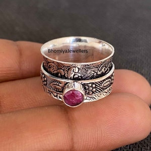 Puede incluir: Un anillo giratorio de plata con un intrincado diseño floral y una piedra preciosa rosa en el centro. El anillo está grabado con el texto "Bhomiyajewellers".