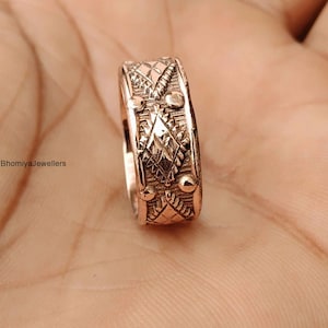 Op de afbeelding: Een ring van roségoudkleurig metaal met een herhalend patroon van gegraveerde ruiten en kleine verhoogde ronde stippen. De band heeft een textuurafwerking met geometrische details over het gehele oppervlak. De tekst BhomiyaJewellers is zichtbaar.