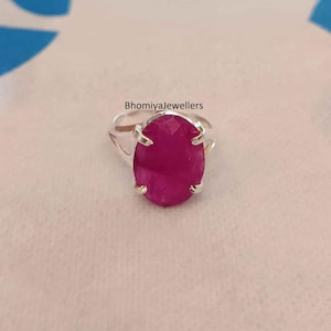 Puede incluir: Un anillo de plata con una gran piedra de rubí rosa de forma ovalada. El anillo tiene un diseño sencillo con cuatro garras que sujetan la piedra en su lugar. El texto "Bhomiyajewellers" es visible sobre el anillo.