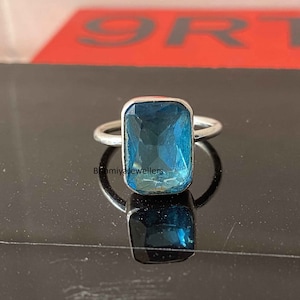 Könnte beinhalten: Ein silberner Ring mit einem großen, rechteckigen, blauen Edelstein. Der Edelstein ist facettiert und hat eine hellblaue Farbe.