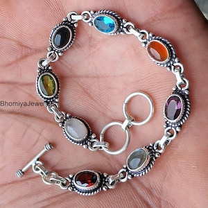 May include: A silver bracelet with oval-shaped gemstones in various colors, including blue, green, red, and purple. The bracelet has a toggle clasp and is designed with a decorative border around each stone. The text "BhomiyaJewels" is visible.