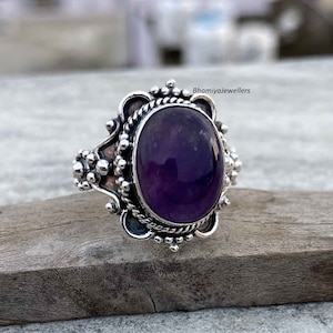 May include: A silver ring with an oval, deep purple gemstone. The ring has an ornate, detailed setting with a textured design around the gemstone and on the band. The ring is set on a wooden surface.