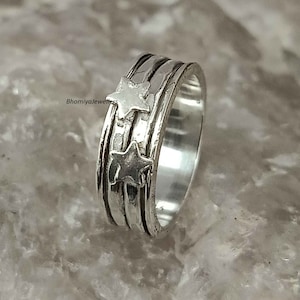 Könnte beinhalten: Ein silberner Drehring mit zwei sternförmigen Designs. Der Ring hat eine strukturierte Band und ist aus Metall.