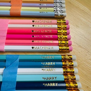 May include: A selection of personalised pencils in pastel shades, each bearing a name in gold lettering. The pencils are bundled with tape. The pencils are ready for use and are in a variety of colours.