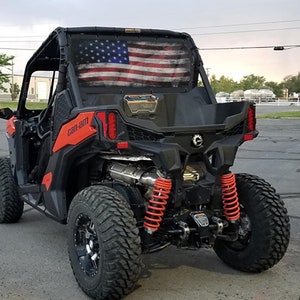 May include: A black Can-Am off-road vehicle with a red frame and a distressed American flag window screen. The vehicle has black wheels with silver rims and red coil springs.