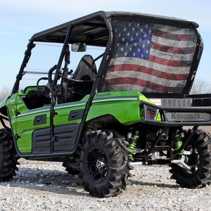 May include: Green Kawasaki utility vehicle with a black roll cage and a black rear bumper. The vehicle has a black and white American flag on the back window. The vehicle is sitting on a gravel road.