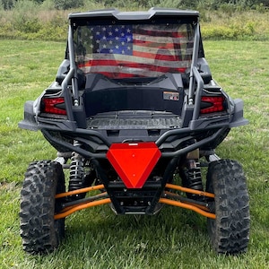 May include: A black off-road vehicle with a red triangle-shaped rear bumper. The vehicle has a rear window with a US flag printed on it. The vehicle is parked on a grassy field.