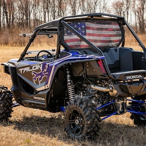 May include: A blue and black Honda Talon side-by-side UTV with large, rugged tires. The vehicle has a roll cage and a rear window featuring an American flag design. The word "TALON" is visible on the side.