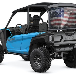 May include: A light blue Yamaha RMAX side-by-side UTV with black accents and large, rugged tires. The rear window features a faded American flag design. The vehicle has a roll cage and the Yamaha logo is visible.