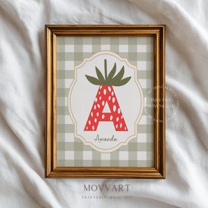 Op de afbeelding: Goudomlijste wandkunst met een rode aardbeivormige letter 'A' met witte stippen, op een groen-witte geruite achtergrond. De naam 'Amanda' staat eronder. De tekst 'MOVART PRINTABLE WALL ART' staat onderaan. Muurdecoratie voor een kinderkamer.