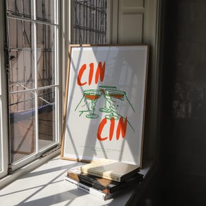 以下が含まれることがあります： オレンジとグリーンの文字「CIN CIN」が書かれた額縁に入ったポスター。ポスターには、ワイングラスを持った4つの手が描かれている。ポスターは白い壁にかけられており、その背後には窓がある。