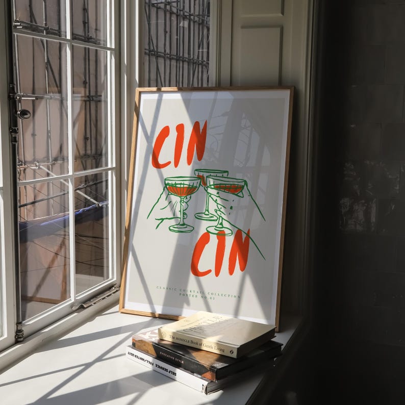 以下が含まれることがあります： オレンジとグリーンの文字「CIN CIN」が書かれた額縁に入ったポスター。ポスターには、ワイングラスを持った4つの手が描かれている。ポスターは白い壁にかけられており、その背後には窓がある。