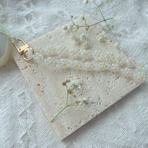 May include: A white beaded keychain with a gold clasp. The keychain is on a light beige textured surface with a small sprig of white flowers.
