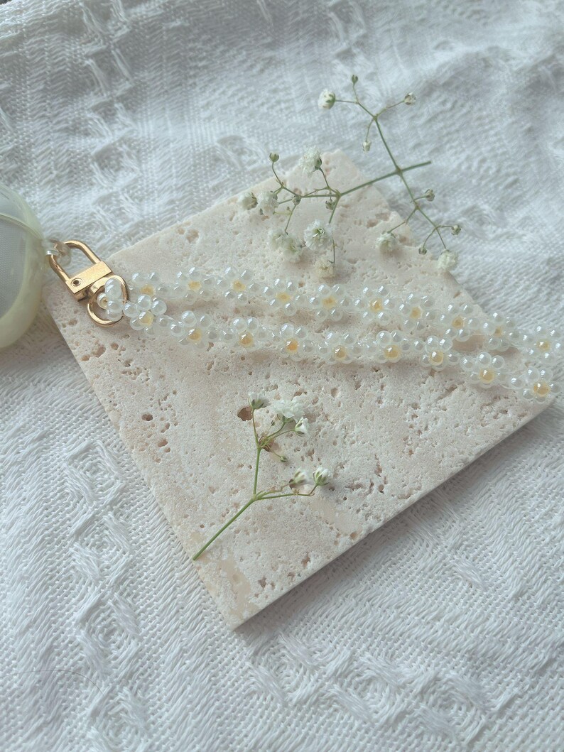 May include: A white beaded keychain with a gold clasp. The keychain is on a light beige textured surface with a small sprig of white flowers.