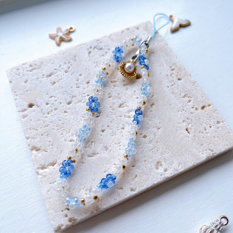 May include: A white and blue beaded phone charm with a gold accent and a small seashell charm. The charm is on a white marble surface.