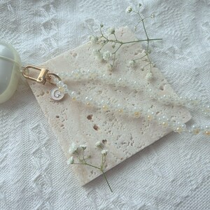 May include: A white beaded phone charm with a gold crescent moon charm attached. The charm is on a white marble surface with a small sprig of white flowers.