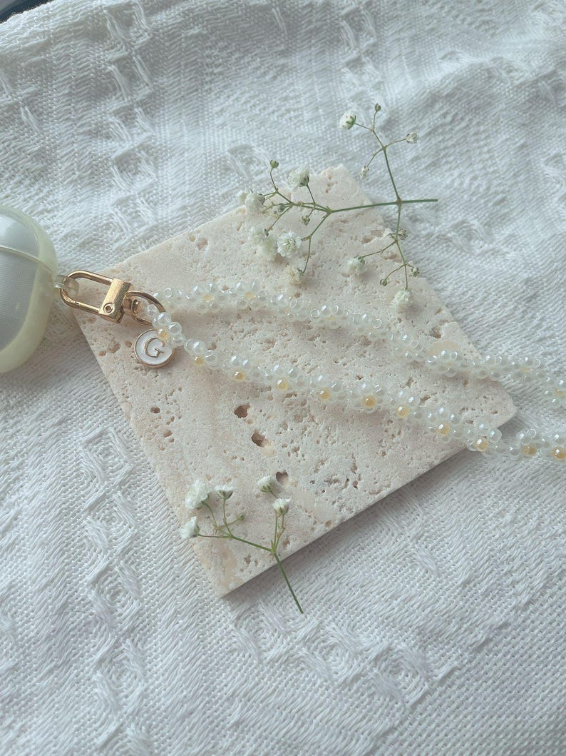 May include: A white beaded phone charm with a gold crescent moon charm attached. The charm is on a white marble surface with a small sprig of white flowers.