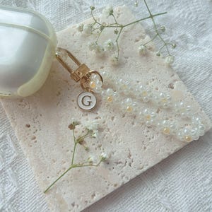 May include: A white Airpod case with a gold charm attached. The charm has a white pearl bead strand and a gold letter 'G' charm.