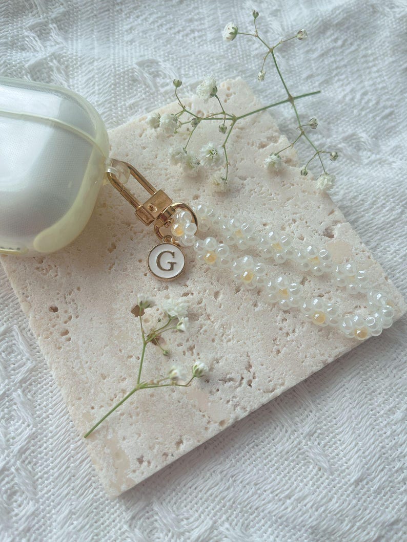 May include: A white Airpod case with a gold charm attached. The charm has a white pearl bead strand and a gold letter 'G' charm.