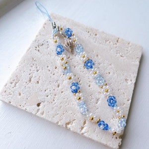 May include: A beaded phone charm with a seashell charm and blue flower accents. The charm is made with white, gold, and blue beads.